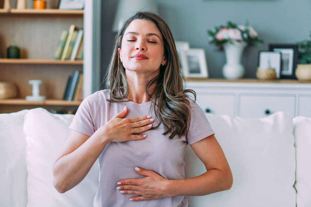 Woman sitting peacefully on a sofa with eyes closed, hands resting on her chest and abdomen in a relaxed breathing posture.